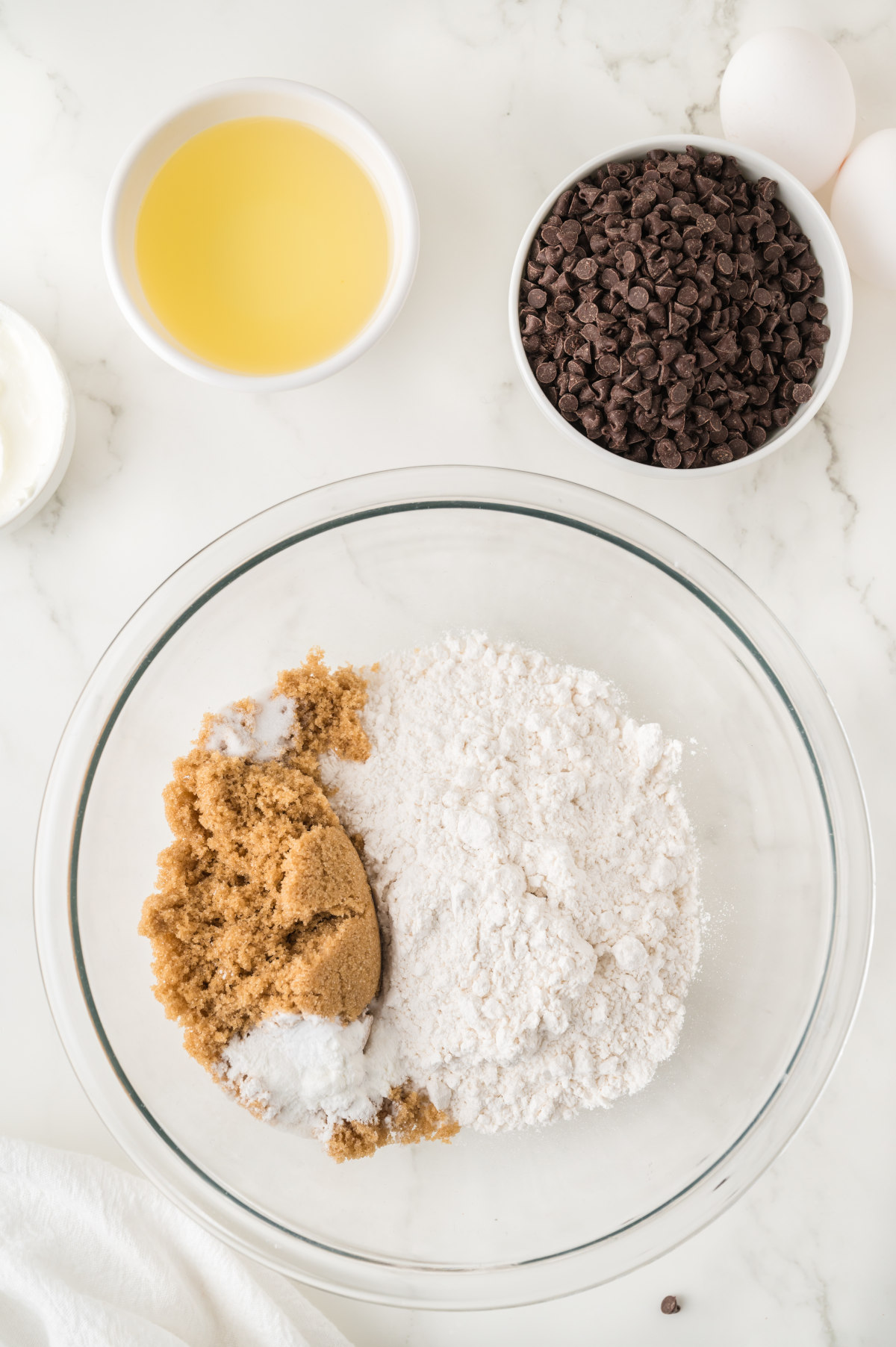 The cake's dry ingredients in a glass bowl, alongside two bowls with the oil and chocolate chips.