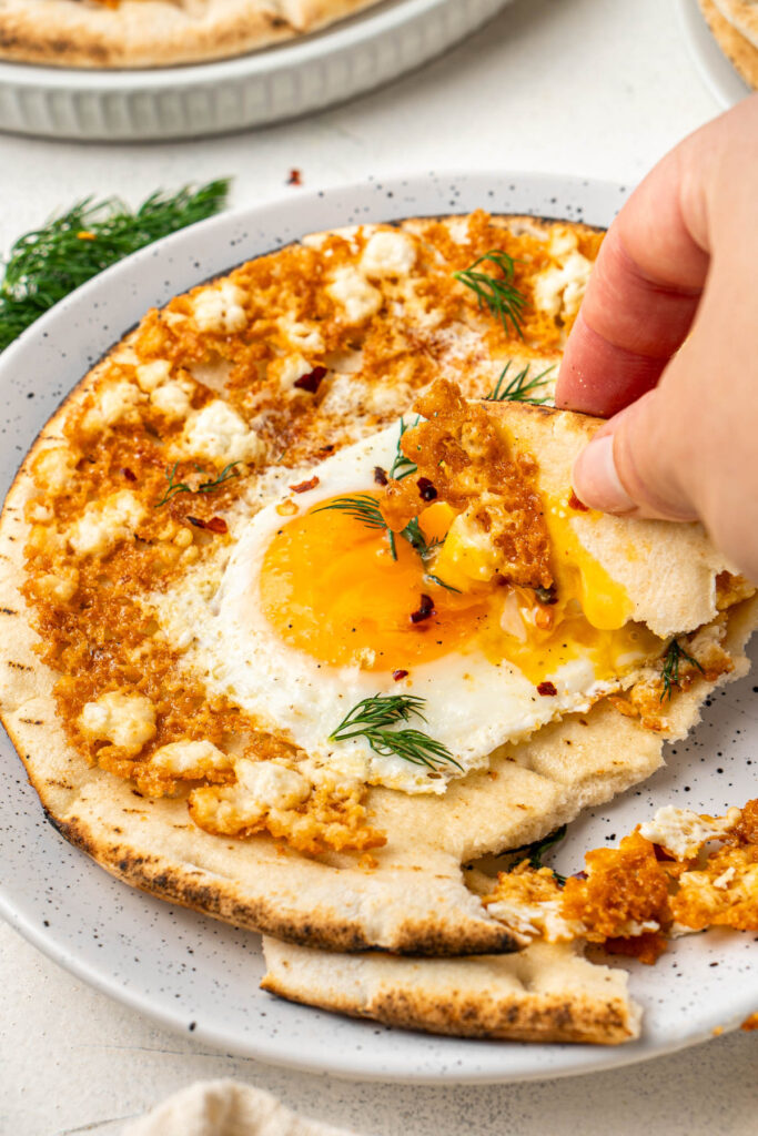A piece of pita bread being dipped into the runny yolk of the finished eggs, served on the rest of the pita bread on a white dish.