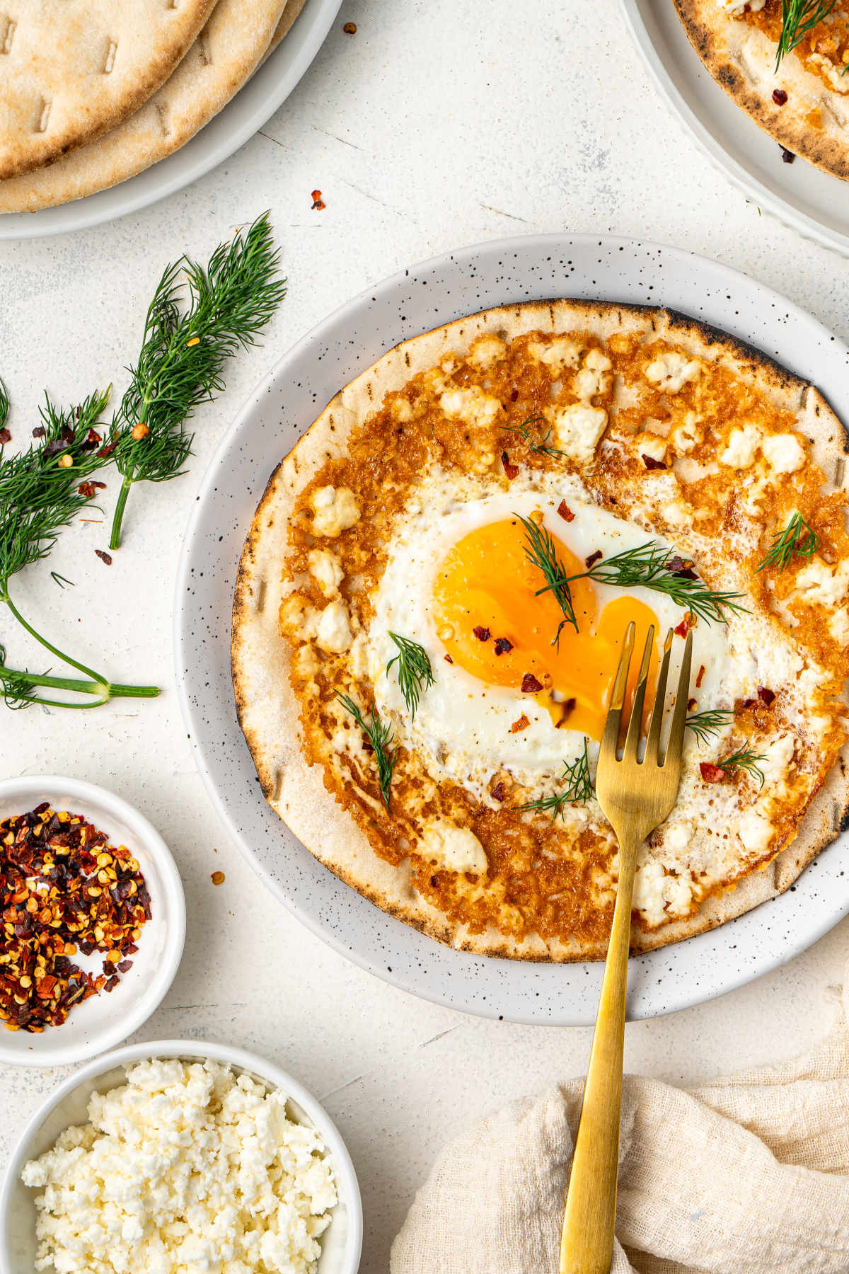 The feta eggs served on a plate with a fork that has broken the yolk and let it run. Its plate is surrounded by its ingredients in white bowls.