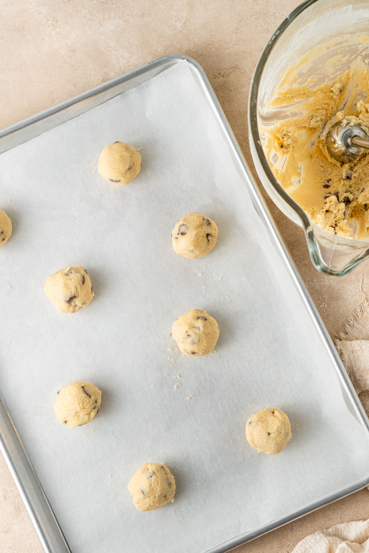 Balls of dough lined up on a baking tray lined with baking paper, beside a glass bowl filled with dough and a teaspoon for making the balls uniform.