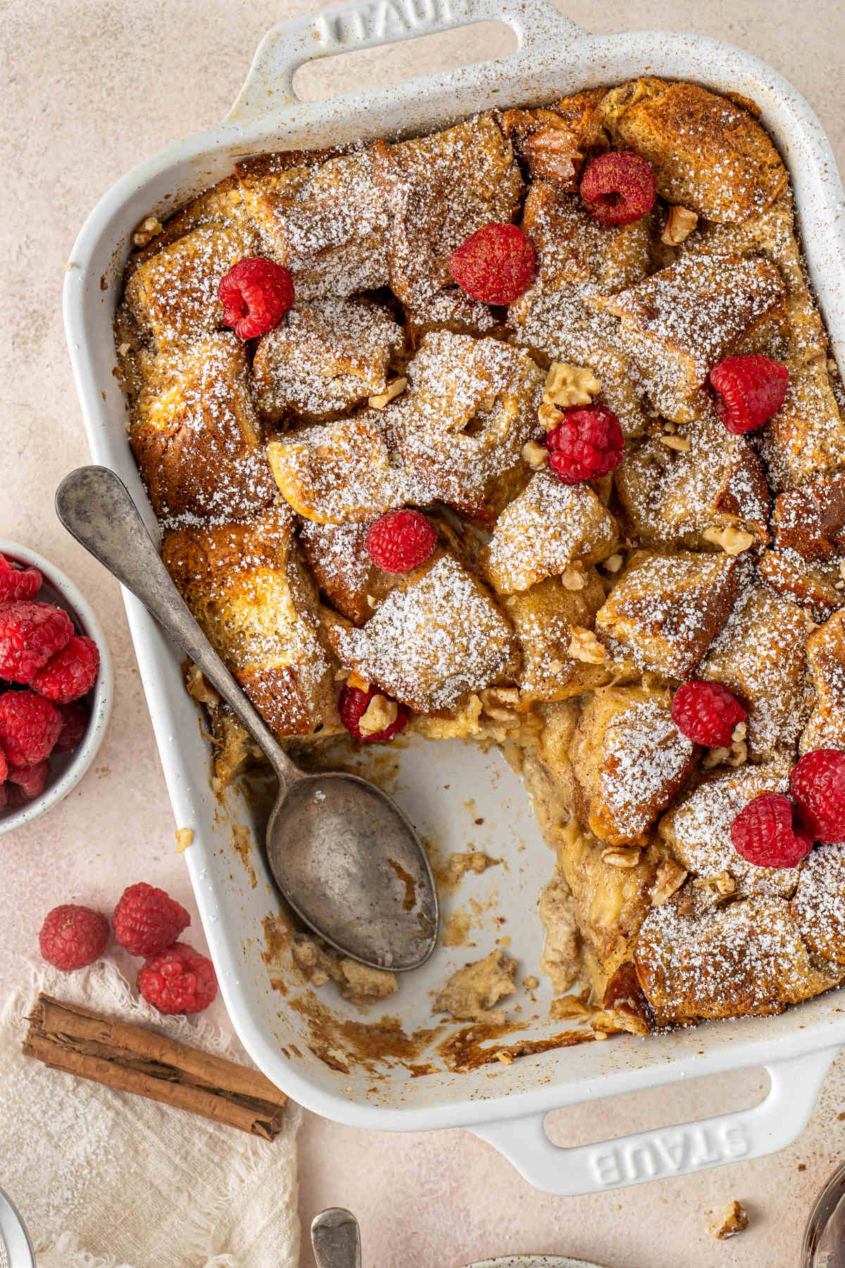 A white baking dish filled with French toast and sprinkled with powdered sugar and raspberries, with a slice taken out and a spoon resting inside of the dish.