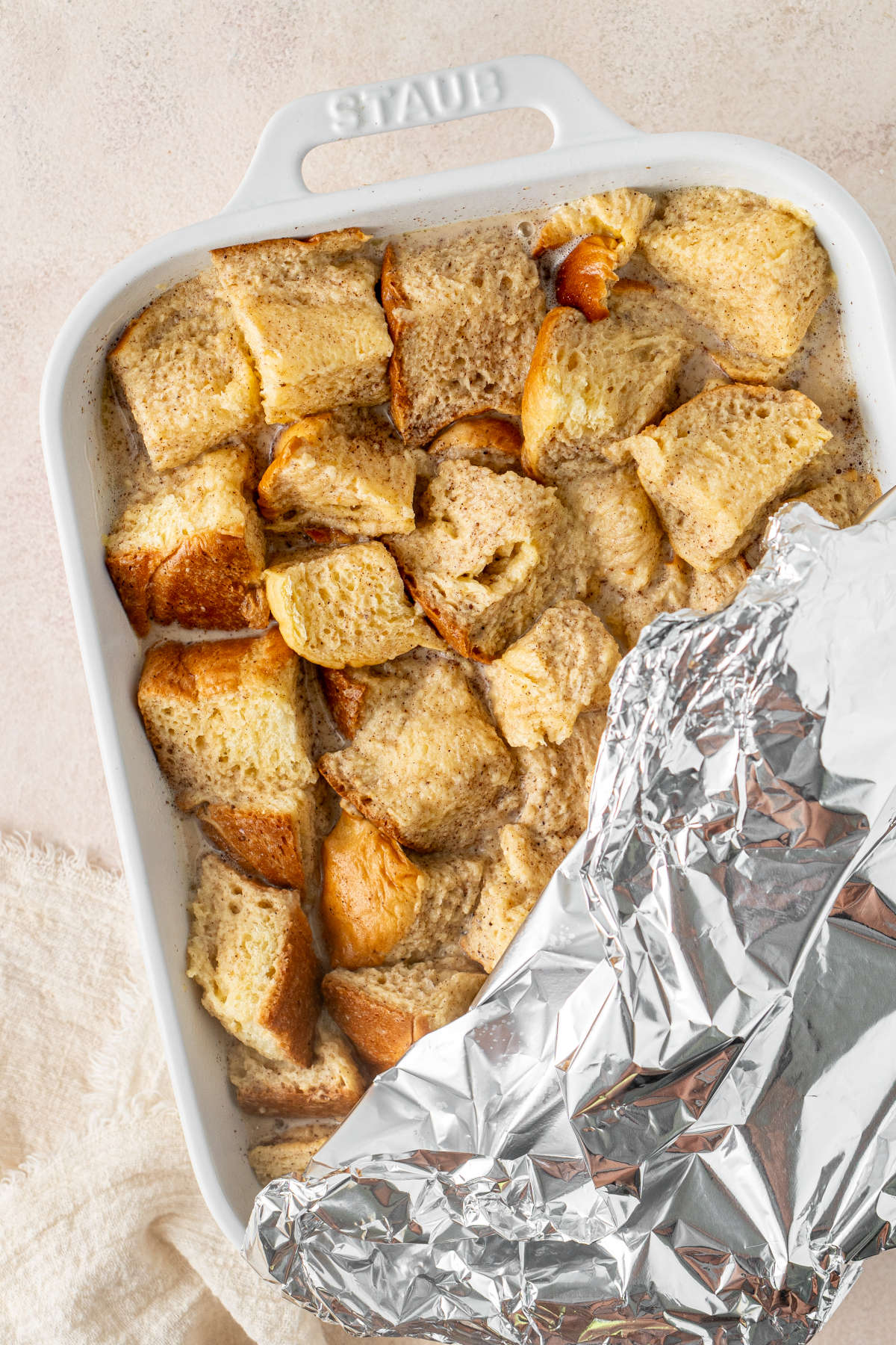 A baking dish full of cubes of brioche soaked in custard, half-covered in a sheet of aluminium foil.