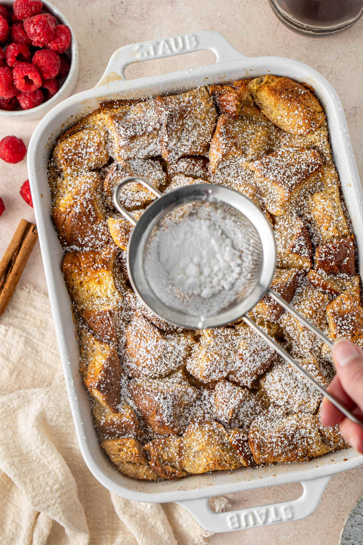 A small sieve of powdered sugar being held over a baking dish of baked French toast which is being dusted with the sugar.