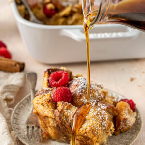 A plate of baked French toast brioche covered in powdered sugar and raspberries being drizzled with maple syrup.