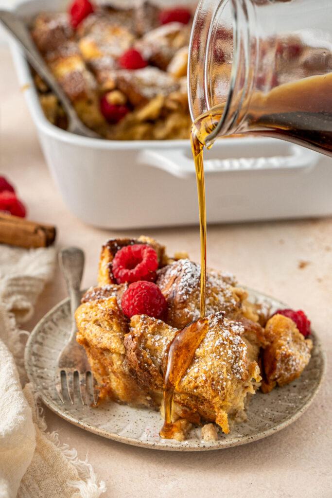 A plate of baked French toast brioche covered in powdered sugar and raspberries being drizzled with maple syrup.