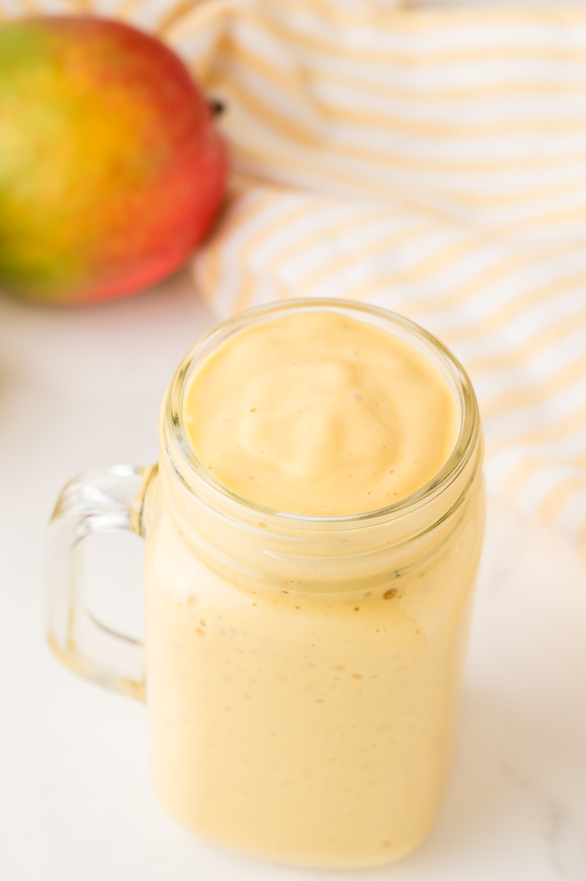 A glass mason jar filled to the brim with milkshake, alongside a mango in the background.