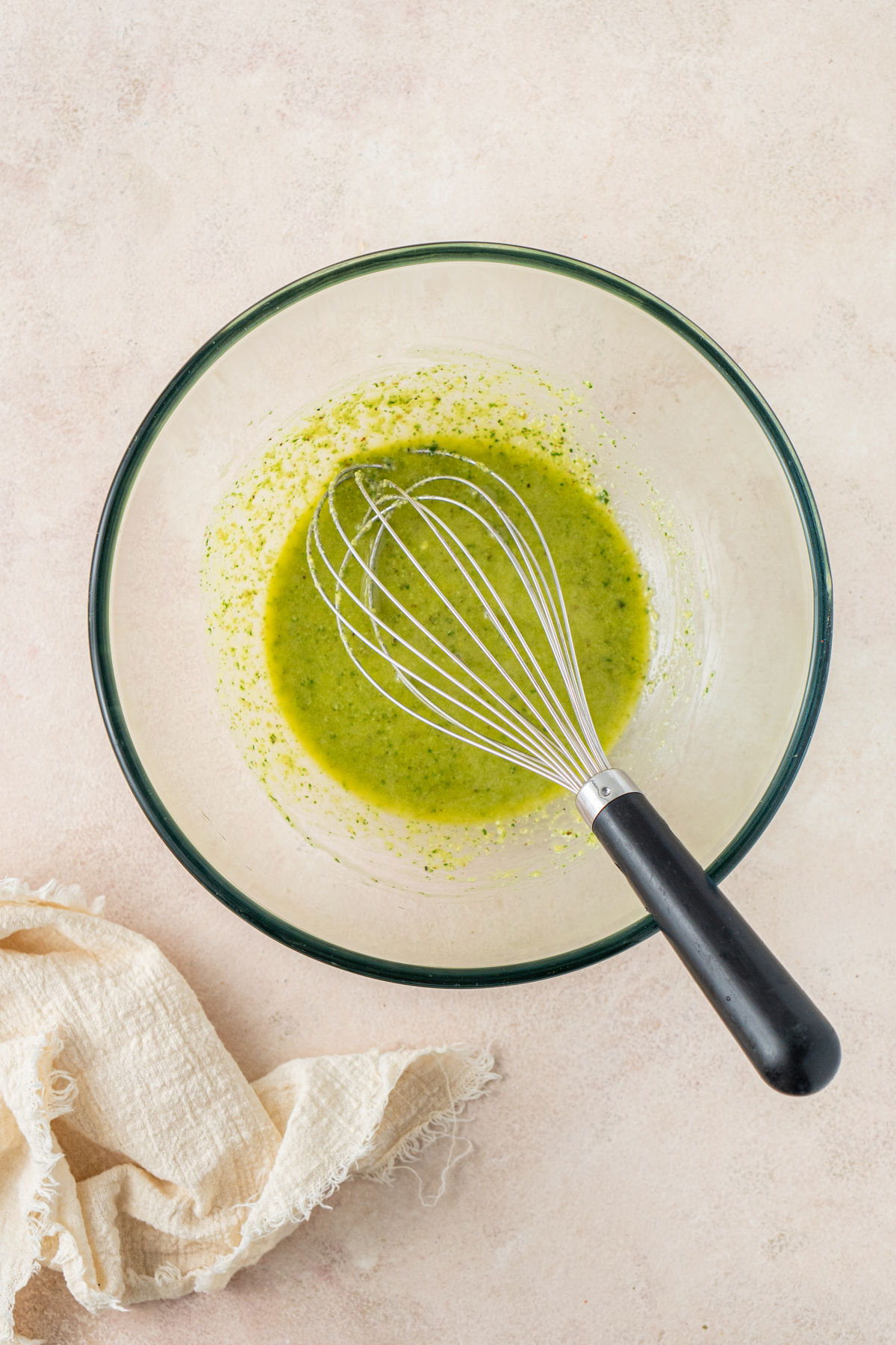 A glass mixing bowl of pesto dressing, with a metal whisk resting in it.