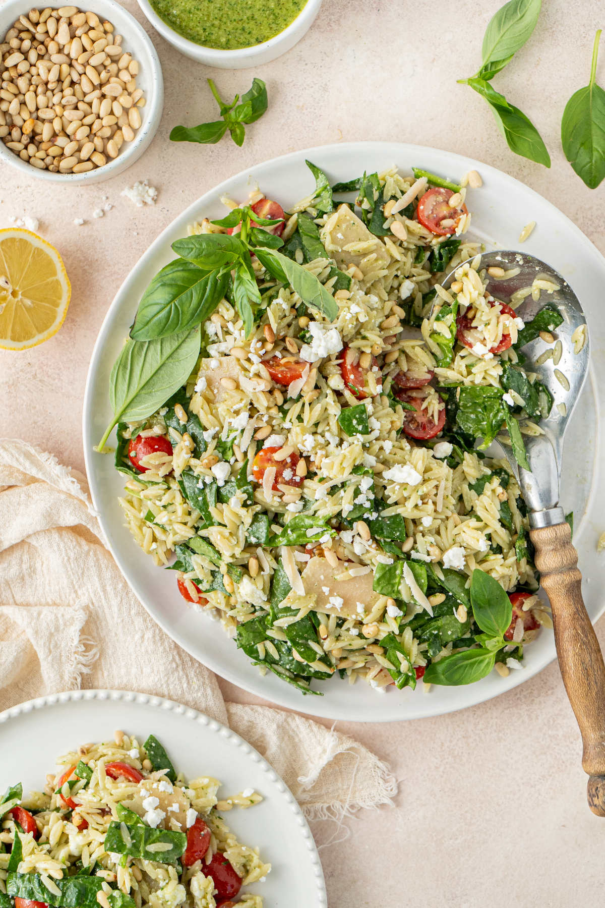 A large plate of salad with a serving spoon resting in it, garnished with basil and surrounded by lemons, pine nuts, and basil leaves.