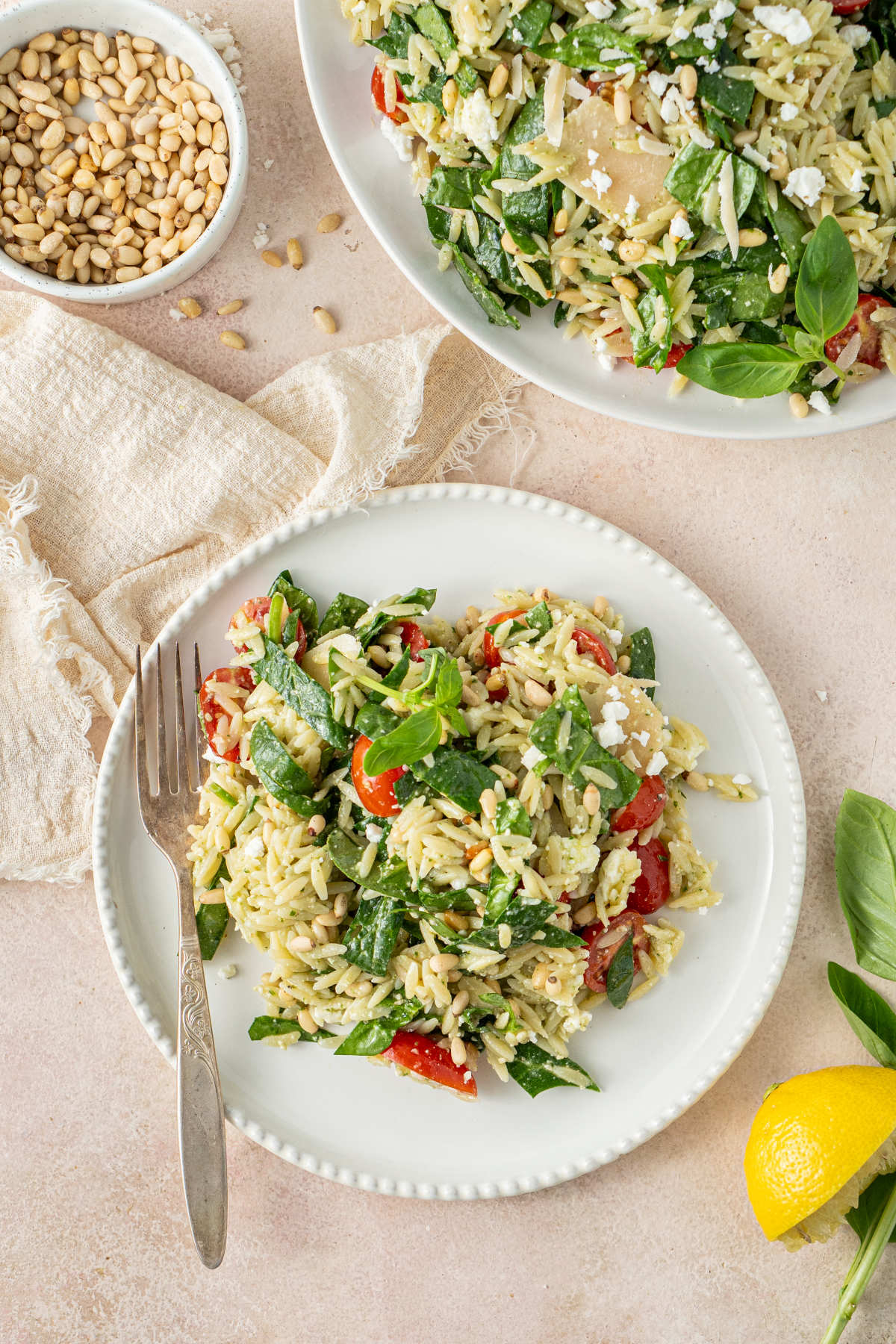 A plate of salad sitting on a kitchen counter, with a fork resting on it and pine nuts and lemons scattered around it, alongside a larger serving bowl.