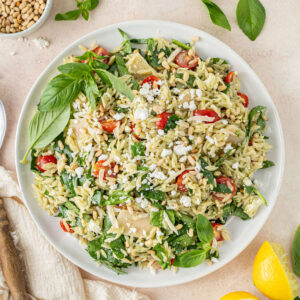 A large serving plate of pesto orzo salad garnished with basil and surrounded by lemons, a bowl of pine nuts, and a bowl of pesto.