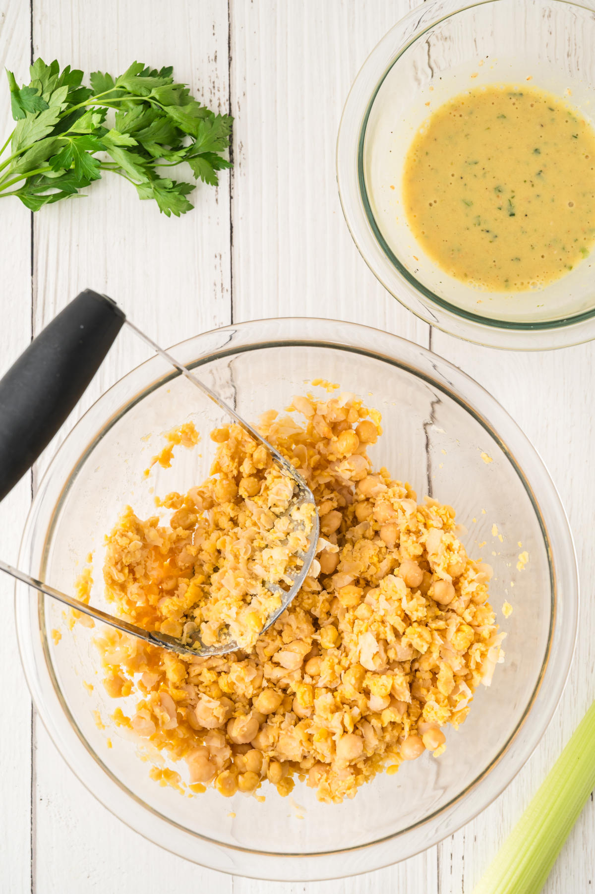 A glass mixing bowl, within which sits half-mashed chickpeas and a potato masher.