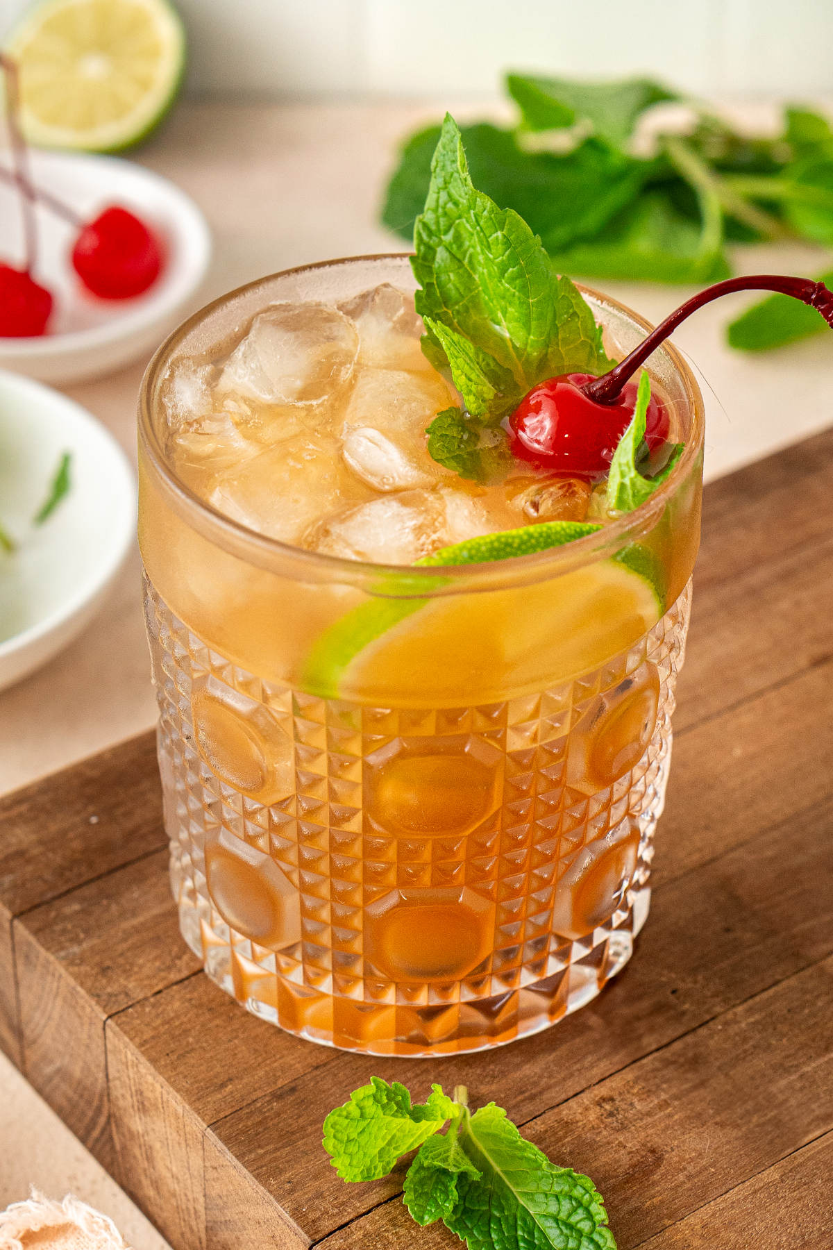A cocktail sitting on a wooden cutting board next to some sprigs of mint and maraschino cherries.
