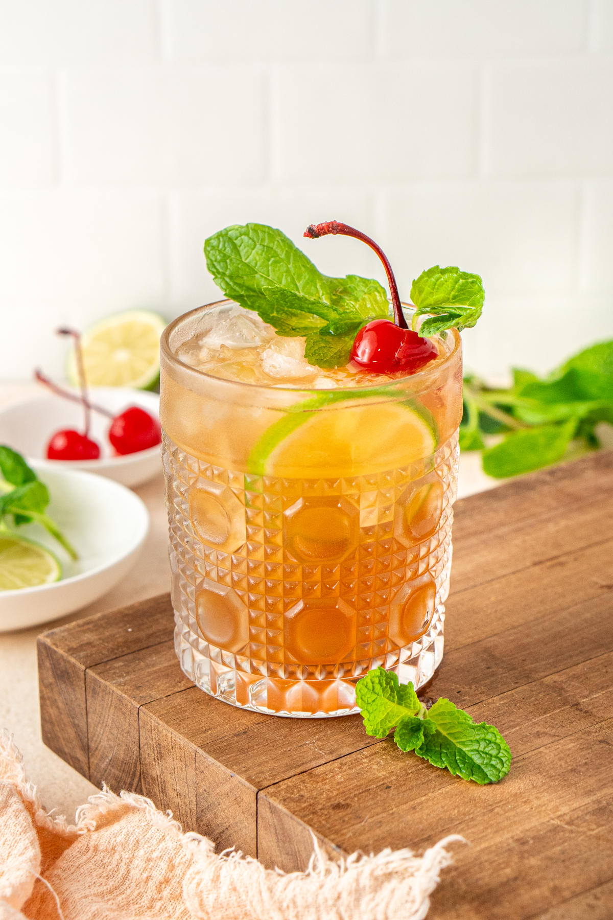 A cocktail sitting on a wooden cutting board next to some sprigs of mint and maraschino cherries.
