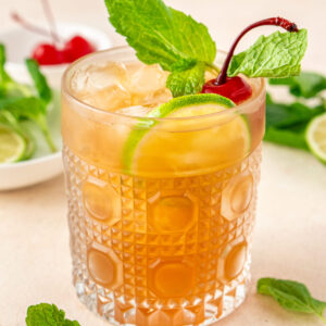 A garnished cocktail in a glass on a kitchen counter surrounded by sprigs of mint.