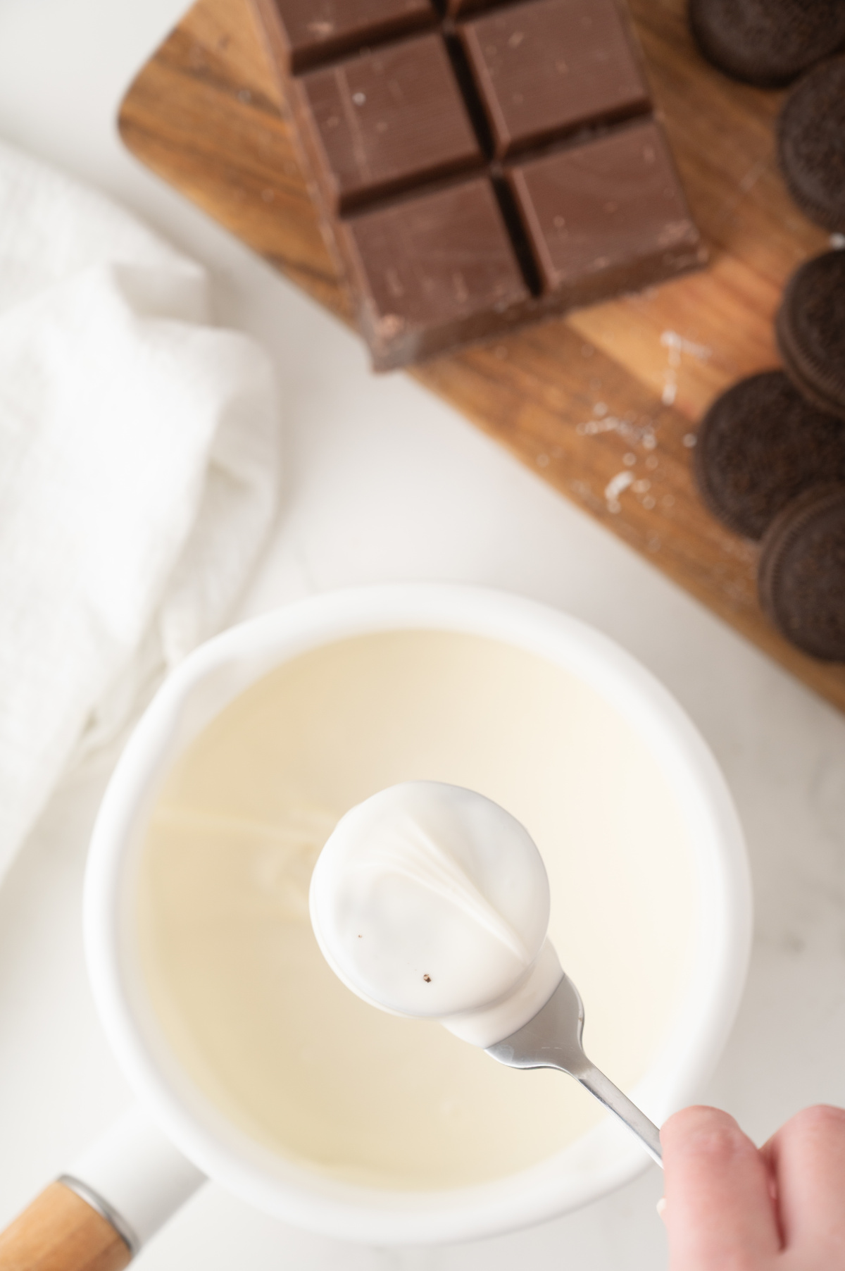 A small saucepan full of vanilla almond bark in the background, with a fork holding a just-dipped Oreo in the foreground.