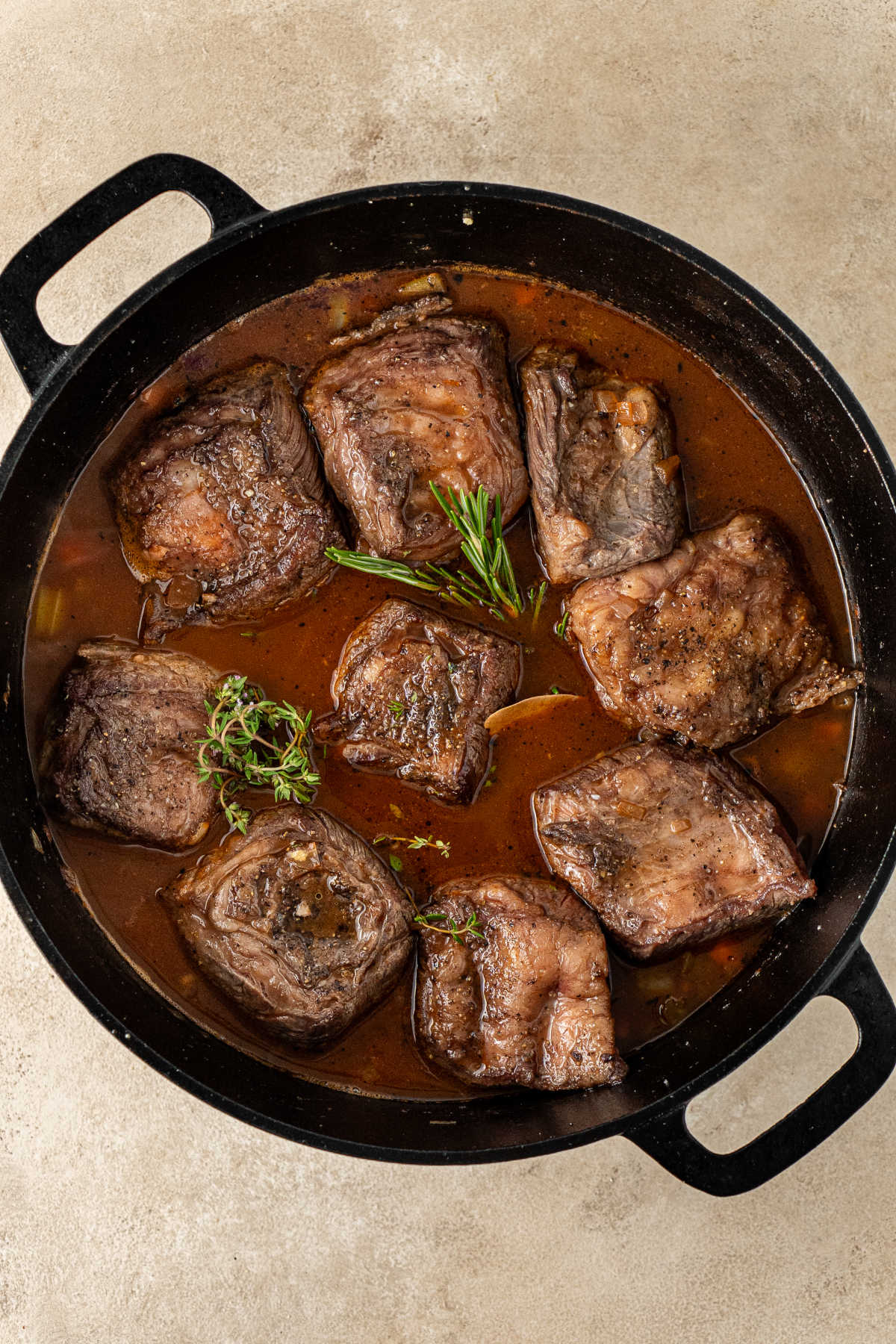 A pot with five seared beef ribs in it, bone side down, halfway-covered in beef broth and sprinkled with herbs.