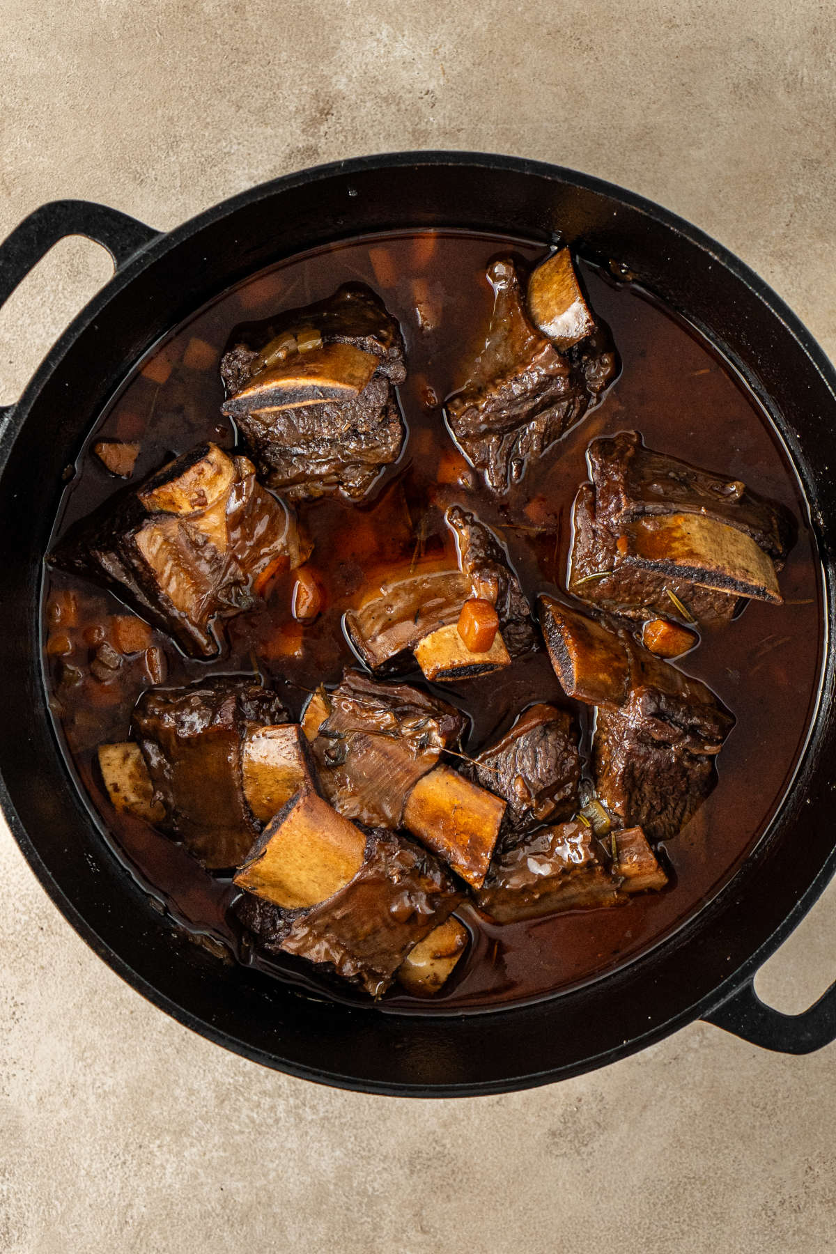 A pot of slow-braised beef, half-covered with a brown broth containing herbs, carrots, etc.