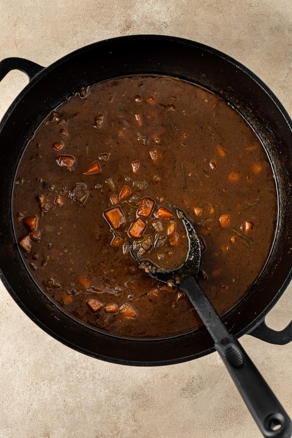 A pot of brown broth containing mirepoix and herbs but without any pieces of beef, within which is resting a black spoon for stirring.