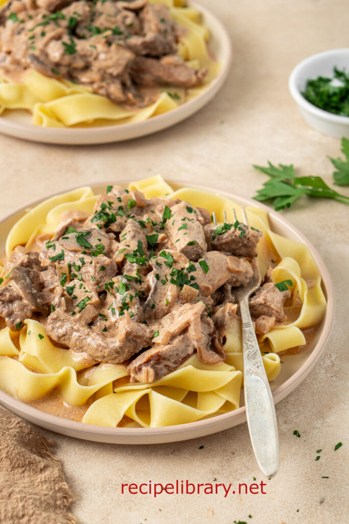 The finished beef stroganoff recipe is served on a plate with pasta. A fork rests on the plate.